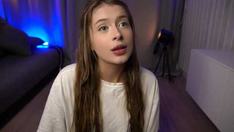 Media: Video of a young Caucasian woman with long brown hair, wearing a white shirt, sitting in a dimly lit room with a blue light, a bed, and a studio light in the background.