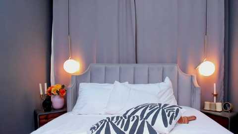 Media: Video of a modern bedroom featuring a light grey tufted headboard, white bedding, a gray patterned blanket, and two gold pendant lamps. The walls are gray, and a vase with flowers sits on a wooden nightstand.