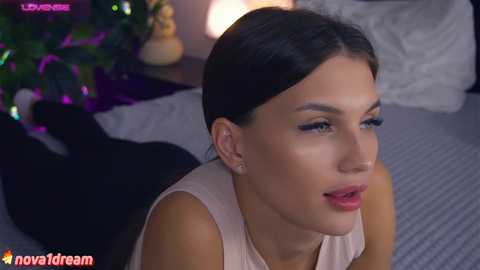 Media: Video of a young woman with dark hair, wearing a beige top, lying on a bed with a black dress. Background includes a Christmas tree, purple lights, and a pillow.