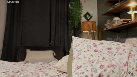 Media: Video of a cozy, dimly lit bedroom with floral-patterned bedding, black curtains, a wooden shelf with a lamp, and a green plant, featuring a soft, warm atmosphere.