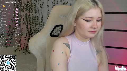 Media: Video of a young woman with fair skin and long blonde hair, wearing a light pink sleeveless top, sitting in a beige gaming chair. Background includes a glittery curtain and a partially visible gaming screen with social media icons.