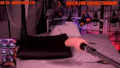 Media: Video of a bedroom with a white bed, black pillow, and a man using a vibrating dildo, surrounded by Halloween decorations and a TV displaying adult content.