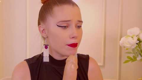 Media: Video of a light-skinned woman with red hair in a bun, wearing black sleeveless top, red lipstick, and long dangling earrings, holding a large, realistic, erect penis in her right hand.