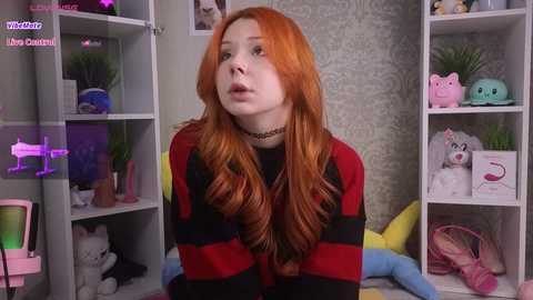 Media: Video of a fair-skinned woman with long, wavy red hair, wearing a striped sweater, in a colorful, plush-filled room with white shelves and a soft gray wall.