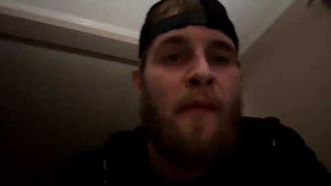 Media: Video of a young man with light skin, a beard, and a black baseball cap, standing in a dimly lit room with a white ceiling and beige wall.