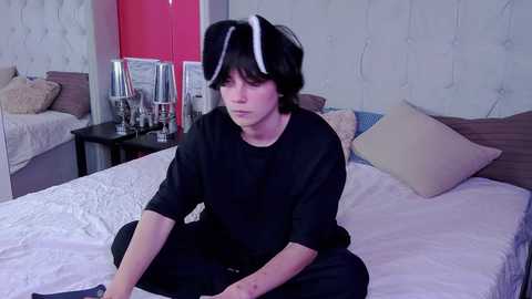 Media: Video of a young person with short black hair, wearing a black T-shirt and white bunny ears, sitting on a bed in a modern, minimalist bedroom with white walls, pink accents, and a wooden headboard.