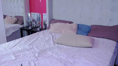 Media: A video of a modern, neatly made bed with white linens, a tufted white headboard, and several pillows in shades of pink, purple, and blue. The background features a mirrored vanity with a red door.