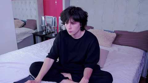 Media: Video of a young, pale-skinned, androgynous person with messy black hair, sitting cross-legged on a white bed in a modern bedroom with a tufted headboard and pink walls.