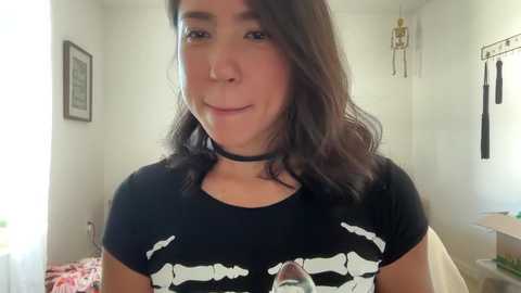 Media: Video of a light-skinned woman with dark hair, wearing a black shirt with white bear prints, and a black choker. Background features a white wall with a skeleton model and a framed picture.