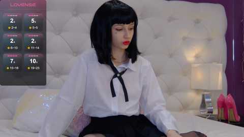 Media: A video of a young woman with straight black hair, red lipstick, and a white blouse with a black bow, sitting on a bed in a plush, white bedroom.