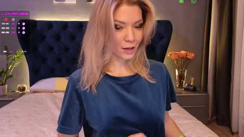 Media: Video of a blonde woman with shoulder-length hair, wearing a blue t-shirt, sitting on a bed with a tufted headboard and floral decorations.