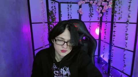 Media: A video of a young woman with pale skin, long black hair, and glasses, sitting in a black gaming chair, surrounded by pink roses and purple lighting.