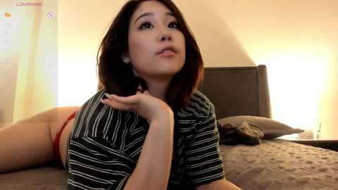 Media: Video of an Asian woman with shoulder-length dark hair, wearing a striped shirt, red panties, lying on a bed with a grey quilt, illuminated by warm bedside lamps.