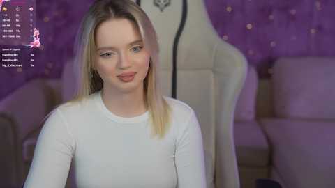 Media: Video of a blonde Caucasian woman with straight hair, wearing a white long-sleeved top, sitting in a gaming chair against a purple wall with bokeh lights. She looks calm and composed.