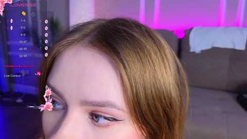 Media: A close-up video of a young woman with fair skin, light brown hair, and striking blue eyes, wearing a pink cherry blossom filter. The background features a pink-lit room with a sofa.