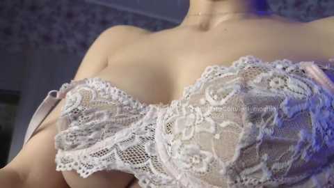 Media: Video of a light-skinned woman with a curvy figure, wearing a lacy, sheer, white bra with intricate floral patterns. Her cleavage is prominently displayed, and the background features a dimly lit, patterned wall.