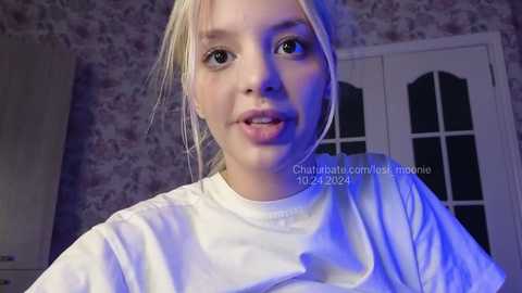 Media: Video of a young woman with light skin and blonde hair, wearing a white T-shirt, standing indoors with floral-patterned wallpaper and a white-framed door in the background.