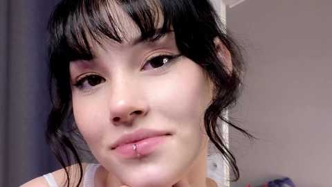 Media: Video of a young woman with light skin, dark hair, and a septum piercing, wearing a white top, against a neutral background.