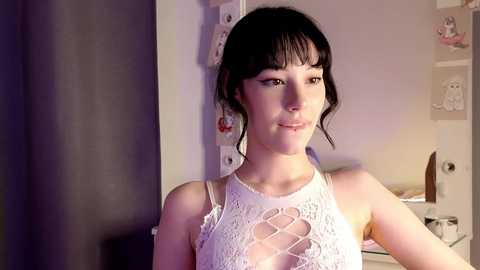 Media: Video of a young woman with short black hair, wearing a white lace top, standing in a dimly lit room with a white door and a cluttered desk.