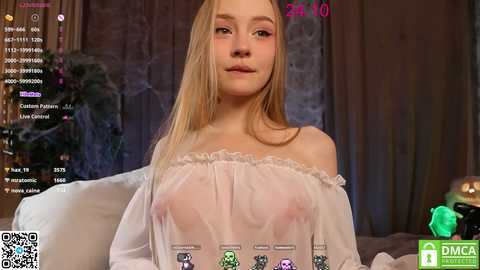 Media: Video of a young, fair-skinned woman with long blonde hair, wearing a sheer, off-the-shoulder top, exposing her small breasts. She's seated in a dimly lit room with a green screen and virtual reality headset, surrounded by gaming elements.