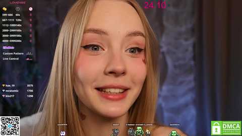 Media: Video of a young, fair-skinned, blonde woman with light makeup, smiling, against a blurred background. Streaming software interface visible with live chat and video settings.