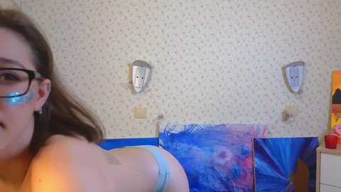 Media: A video of a young woman with long brown hair, wearing glasses, and light blue underwear, lying on a blue tarp in a cluttered, wallpapered room with a TV and wall art.