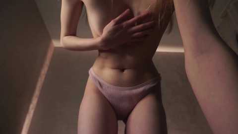 Media: A video of a topless, slender woman with fair skin, wearing pink ribbed panties, standing in a dimly lit hallway with beige walls. Her left hand covers her breast, and her long blonde hair cascades down her back.