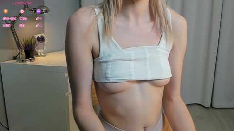 Media: Video of a slender, fair-skinned woman with blonde hair, wearing a white crop top that exposes her small breasts. Background includes a modern desk with decorative items and grey curtains.