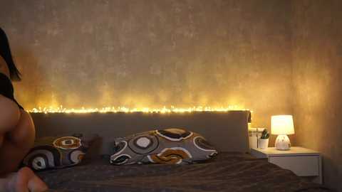 Media: Video of a modern bedroom with a gray upholstered headboard, patterned pillows, beige textured walls, a cozy bedside lamp, and a warm string of fairy lights above the bed.