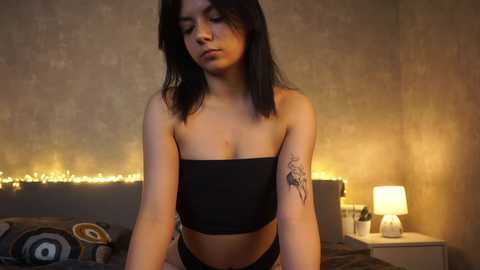 Media: Video of a young woman with medium-length black hair, wearing a black strapless crop top and black panties, standing in a dimly lit bedroom with a beige textured wall, a bed, and a lamp.