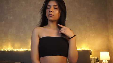 Media: Video of a young woman with long black hair, wearing a black bandeau top, standing against a dimly lit, textured wall with fairy lights. She has a relaxed expression, one hand touching her neck.