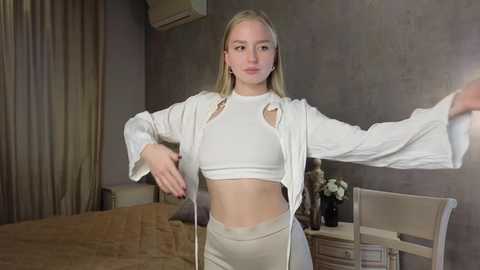 Media: Video of a young, slender, fair-skinned woman with long blonde hair, wearing a white crop top and pants, posing in a modern, minimalist bedroom.