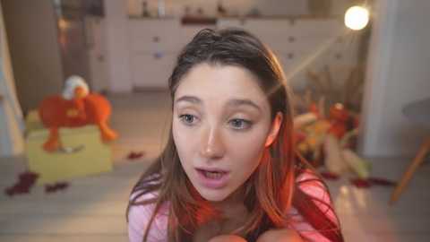 Media: Video of a young woman with fair skin, brown hair, and a surprised expression, kneeling in a messy room with orange and yellow toys scattered around.