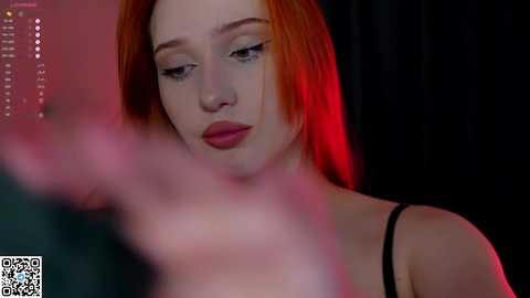 Media: Video of a fair-skinned, red-haired woman with a contemplative expression, wearing a black spaghetti-strap top, against a dark background with red lighting.