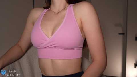 Media: Video of a light-skinned woman with medium-length brown hair, wearing a pink sports bra and black pants, standing in a dimly lit room with a closed door and white towel.