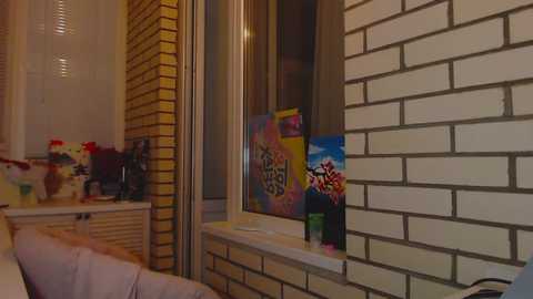 Media: Video of a cozy, well-lit room with a white brick wall, yellow brick accent, a window with a colorful poster, and a small table with a vase of flowers.