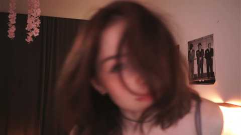 Media: A blurry video of a young woman with shoulder-length brown hair, wearing a black dress, in a dimly lit room with pink cherry blossom branches and a framed black-and-white photo of four people on a white wall.