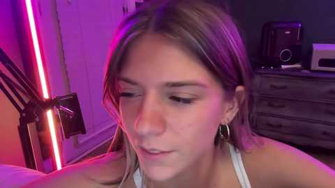 Media: Video of a young woman with light brown hair, wearing a white top, looking down, in a dimly lit room with purple and pink LED lights, a desk, and a speaker.