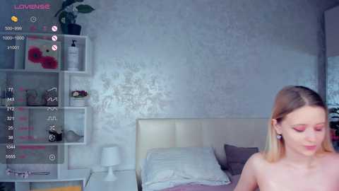 Media: Video of a young, nude, fair-skinned blonde woman with medium-sized breasts, in a modern, white bedroom with a floral-patterned wall, a bed, and a plant.