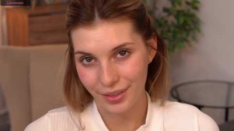 Media: Video of a young, fair-skinned woman with shoulder-length, straight, light brown hair, wearing a white ribbed sweater. She has a slight smile and is indoors with a blurred background featuring a wooden cabinet and potted plant.