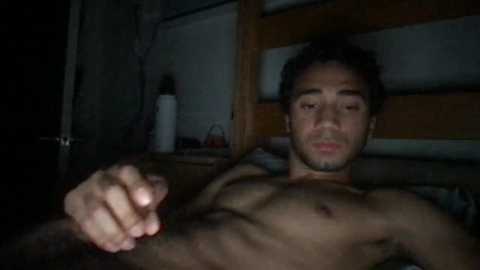 Media: A dimly lit video of a shirtless, muscular man with short dark hair, lying on a wooden bed, making a fist with his right hand, against a shadowy, cluttered background.