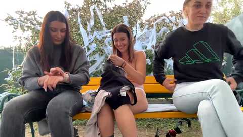 Media: Video of three young women sitting on a yellow park bench, laughing. One is topless, wearing only black shorts, another in a gray sweater, and the third in a black Adidas sweatshirt. Background features a graffiti wall and green trees.