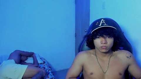 Media: Video of a shirtless young man with curly hair and a black A helmet, sitting indoors. Blue lighting casts a cool hue.