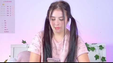 Media: A young woman with long black pigtails, fair skin, and a pink t-shirt, sits in a softly lit room with green ivy, a calendar, and a phone.