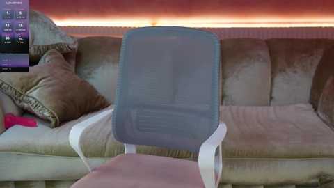 Media: Video of a beige velvet couch with a light blue mesh chair in front. Background has a radiant orange glow and a pink cushion on the left.