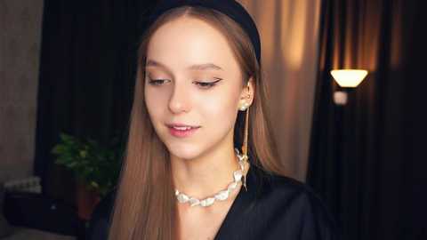 Media: Video of a young woman with fair skin, long straight brown hair, wearing a black headband, black dress, and silver necklace. Background features dim lighting, a potted plant, and a lamp.