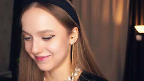 Media: Video of a young woman with light skin and straight brown hair, adorned with a black headband, wearing a black dress and long, dangling gold earrings. She has a subtle smile, with a warm, blurred background.