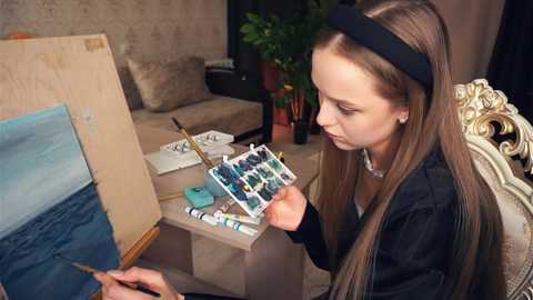 Media: Video of a young girl in a black headband painting on a wooden easel in a cozy living room with beige furniture.