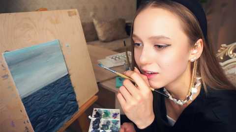 Media: Video of a young girl with light skin, straight brown hair, wearing a black beret and necklace, painting a seascape on an easel in a studio with beige walls.