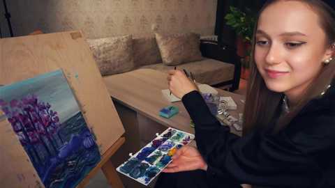 Media: Video of a young woman with long brown hair, wearing a black jacket, painting an underwater scene on a wooden easel in a cozy room with beige walls and a potted plant.